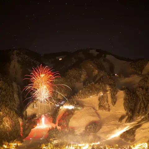 Fireworks light up the night sky over a snowy mountain resort, with bright ski trails and a starry sky in the background