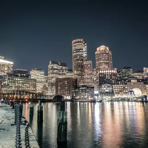 Boston skyline at night, with illuminated skyscrapers reflecting on the calm waters of the harbor.