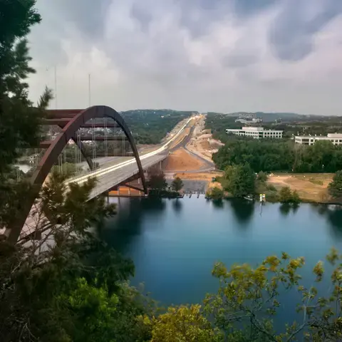 Pennybacker Bridge in Austin, Texas, spanning the Colorado River, with surrounding greenery and distant office buildings