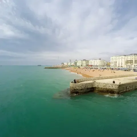 Coastal view with turquoise water, a stone pier, and a sandy beach lined with classic white buildings under a cloudy sky