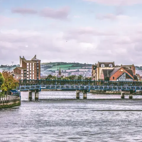 Brick buildings line the riverside with a blue pedestrian bridge spanning the water and rolling green hills in the background