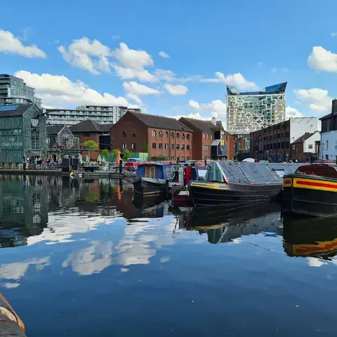 Canal boats docked on calm water, surrounded by historic and modern buildings under a bright blue sky with clouds